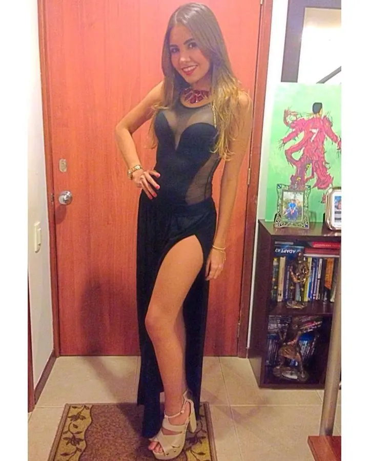 Jeune femme latine en robe noire fendue, posant près d'une porte, souriant à l'appareil