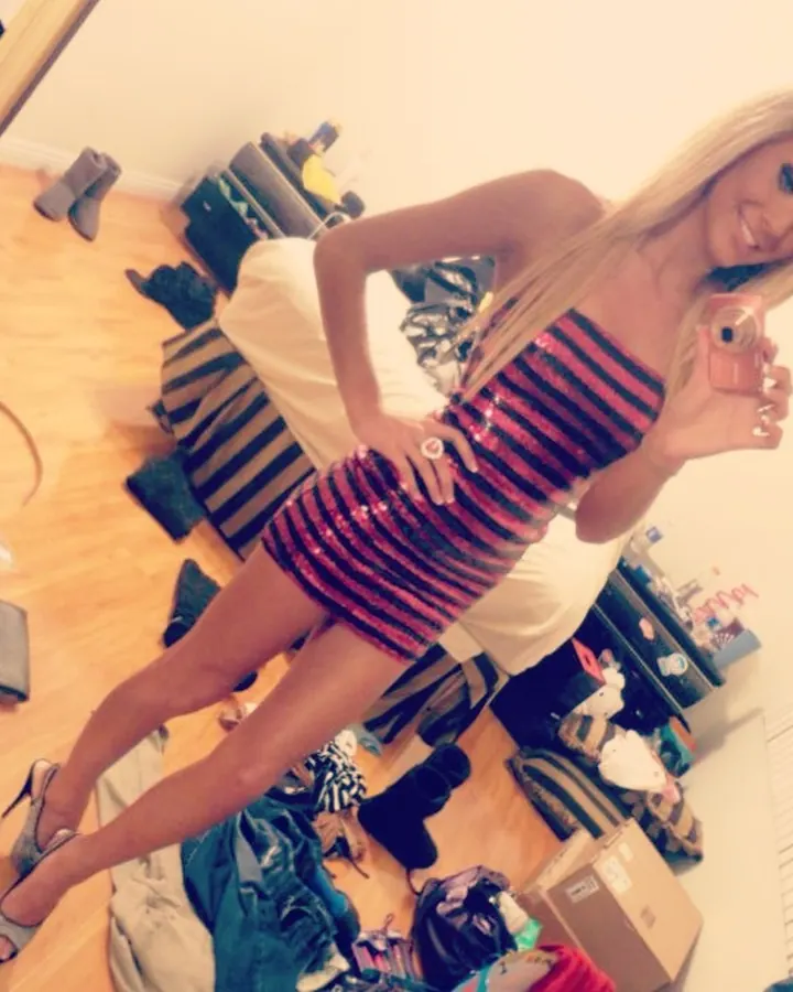 Jeune femme blonde aux cheveux longs, portant une robe à rayures roses et noires scintillantes, prenant un selfie dans une chambre en désordre, entourée de chaussures et de vêtements.