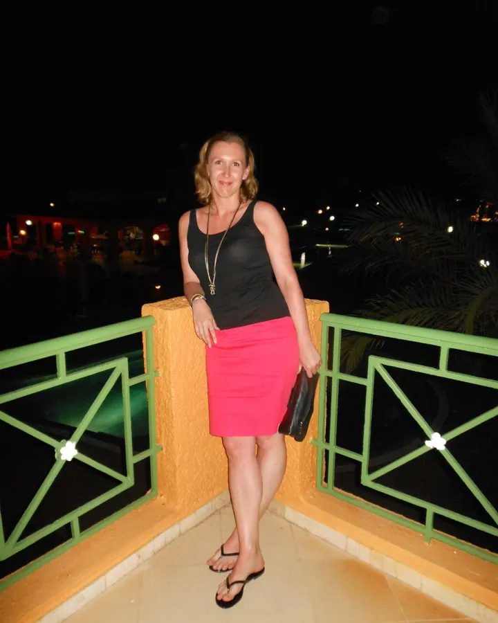 Femme souriante en tenue décontractée (débardeur noir, jupe rose) sur une terrasse nocturne, avec vue sur des palmiers et des lumières lointaines.