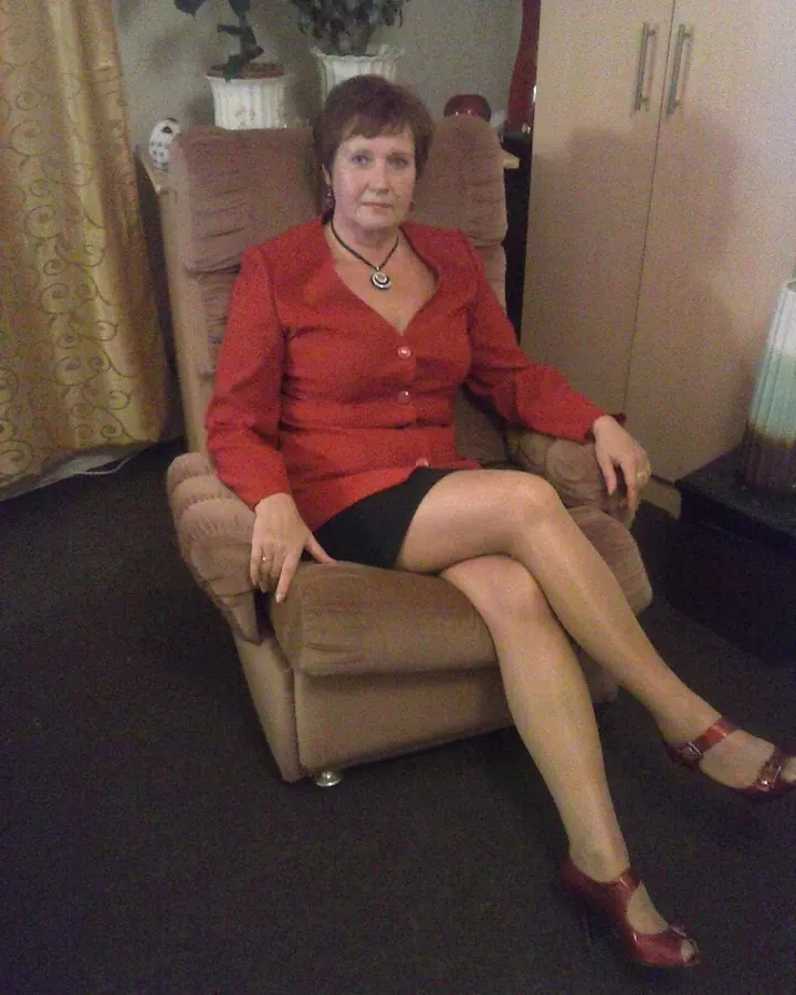 Femme aux cheveux châtains portant une robe rouge, assise dans un fauteuil, les jambes croisées, dans un salon sombre.