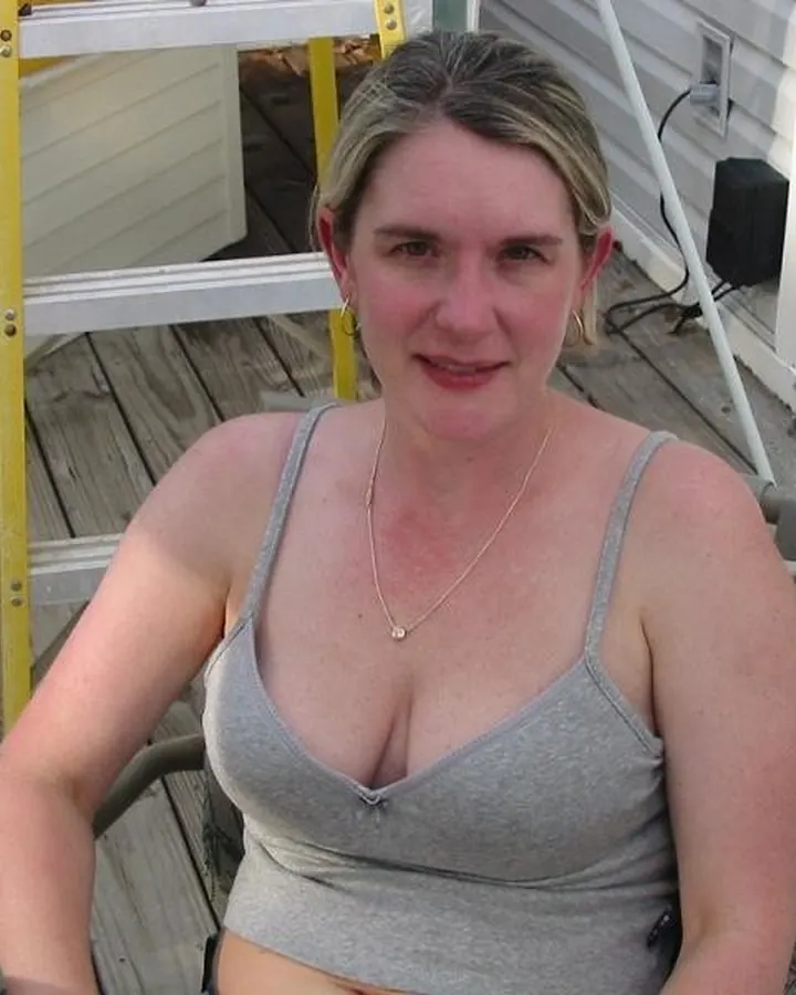 Femme aux cheveux blonds, assise sur une terrasse en bois, portant un haut gris sans manches, avec un petit collier doré.