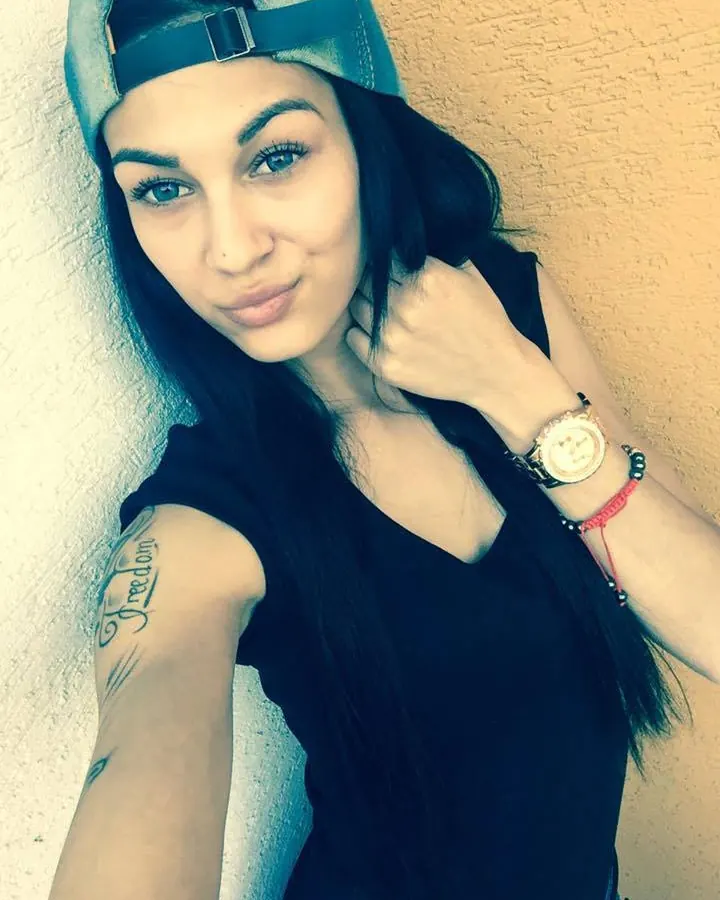 Jeune femme aux cheveux foncés, casquette renversée, regardant la caméra, tatouage visible sur le bras.
