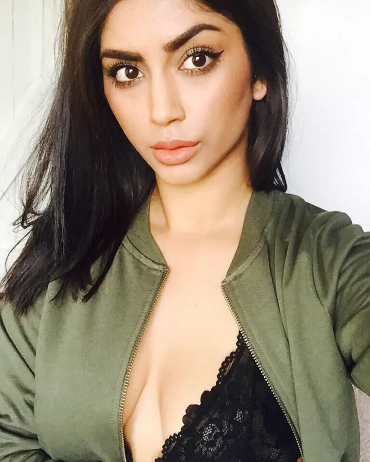 Femme aux cheveux bruns, portant un haut vert et une jupe courte frangée, debout sur un fond gris.
