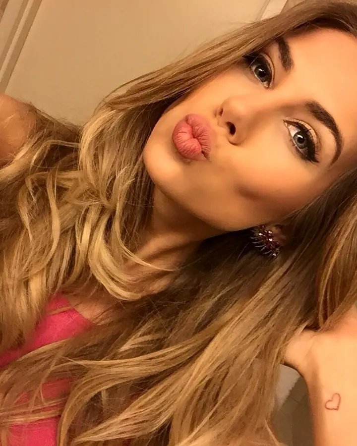 Femme blonde aux cheveux longs ondulés, portant un haut rose, faisant un baiser à la caméra, un petit cœur tatoué sur le bras.