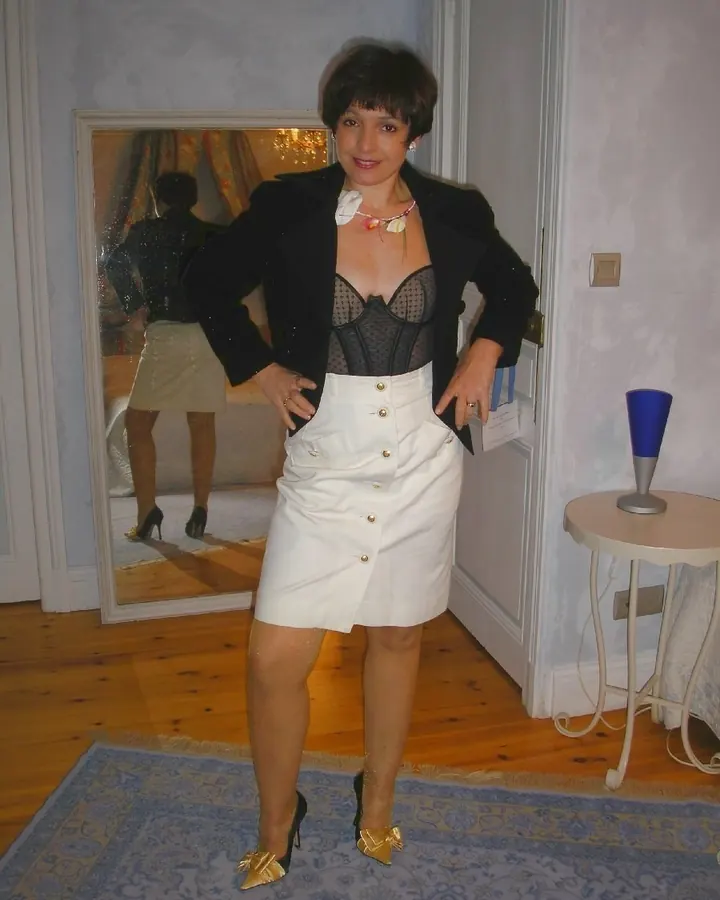Femme aux cheveux bruns courts, en blazer noir et corset noir, jupe blanche boutonnée, chaussures dorées, posant devant un miroir.