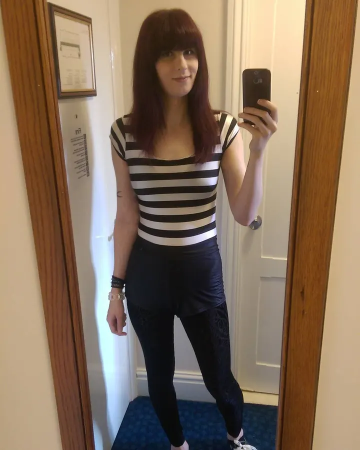 Femme aux cheveux châtains, en bikini noir, posant devant un miroir, tenant un téléphone rose.