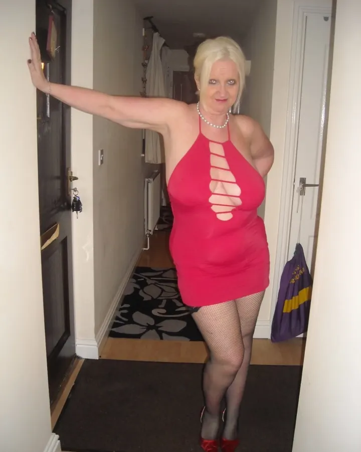 Femme blonde en robe rouge fendue, bas résille et escarpins rouges, posant dans un couloir