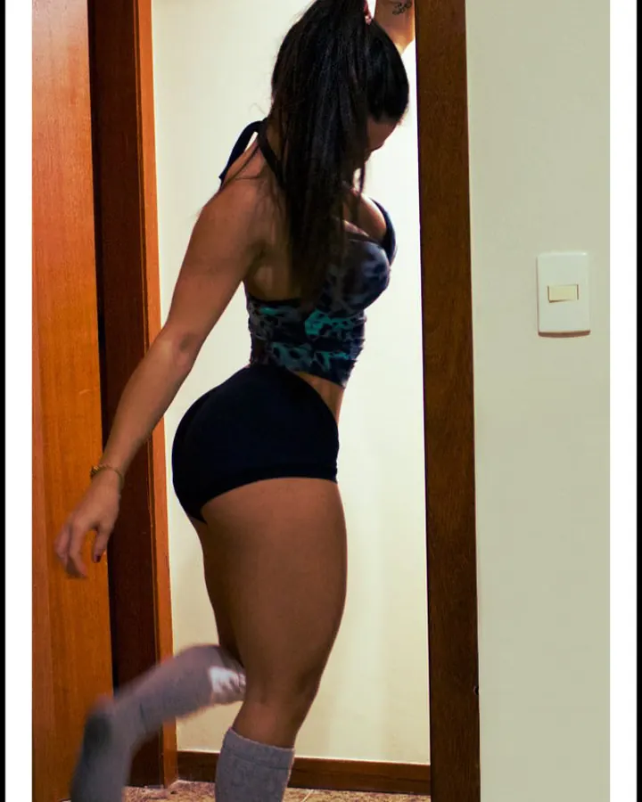Jeune femme latino aux cheveux châtains, vêtue d'un haut noir, d'un short blanc et de collants gris, en train de marcher dans un couloir, les jambes gainées et les chevilles montées.