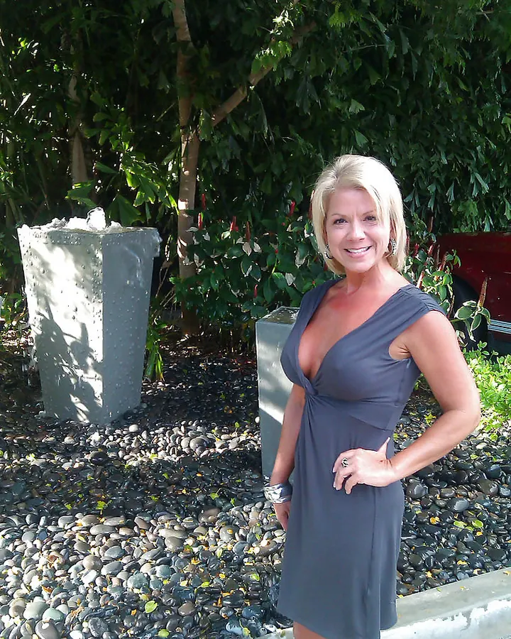 Femme blonde en robe grise ajustée, debout sur un chemin pavé, devant des plantes vertes, main sur la hanche, souriant à l'appareil.