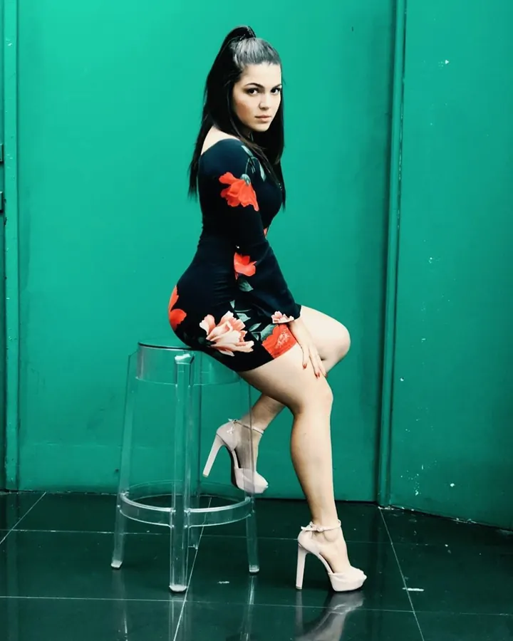 Femme aux cheveux noirs en queue-de-cheval, en robe noire à fleurs rouges, assise sur un tabouret transparent devant un mur vert.