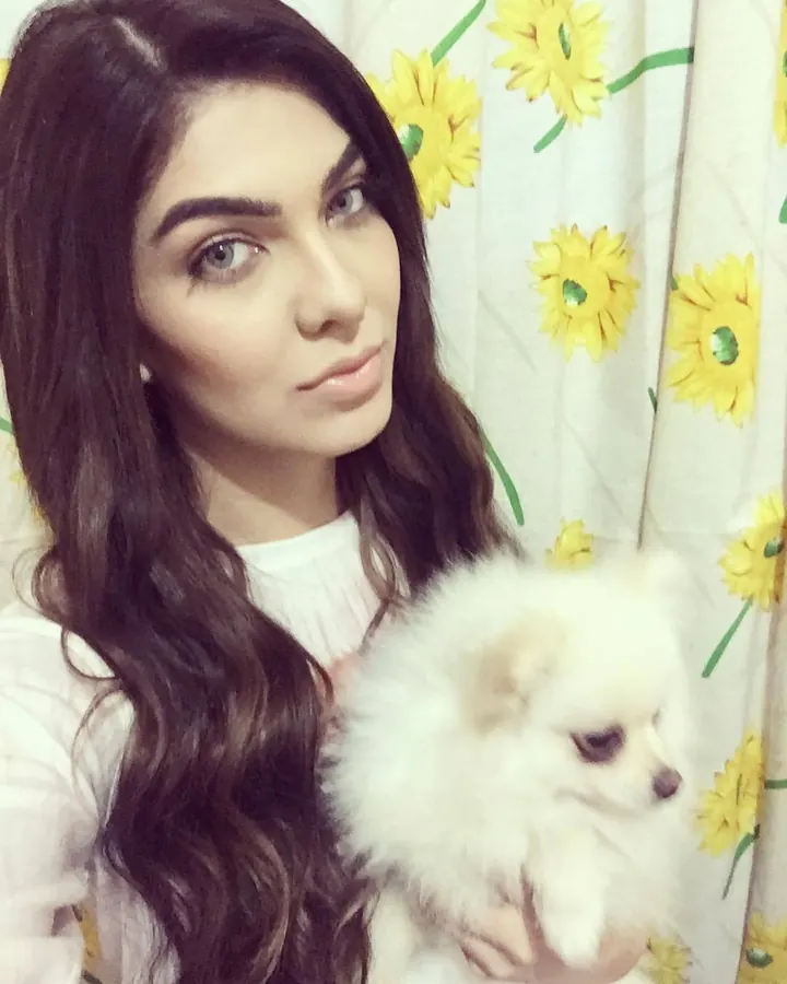 Femme aux cheveux bruns, tenant un petit chien blanc, devant un rideau à fleurs jaunes.