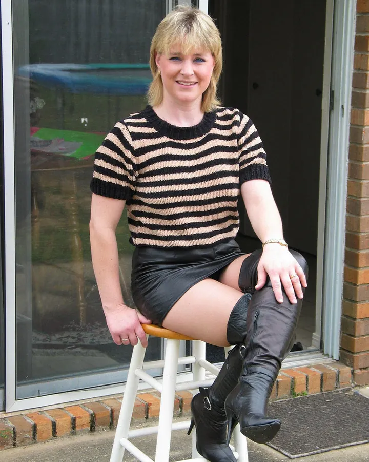 Femme blonde assise sur un tabouret, en pull rayé noir-beige, jupe en cuir noir et bottes hautes noires, souriante.