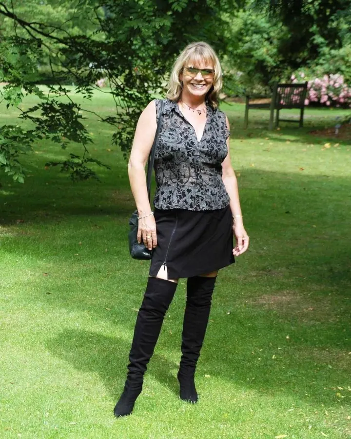 Femme blonde en tenue sombre, debout dans un jardin, portant des lunettes, souriante.