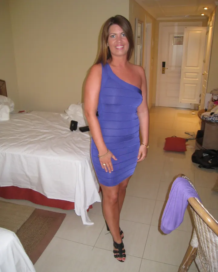 Femme blonde en robe bleue ajustée, souriante, debout dans une chambre d'hôtel.