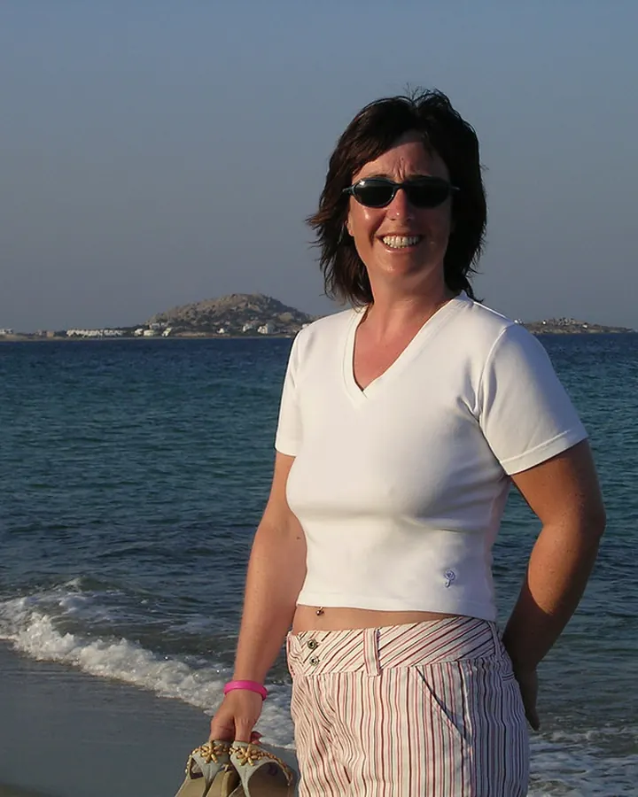 Femme brune en t-shirt blanc, souriante, debout sur une plage.