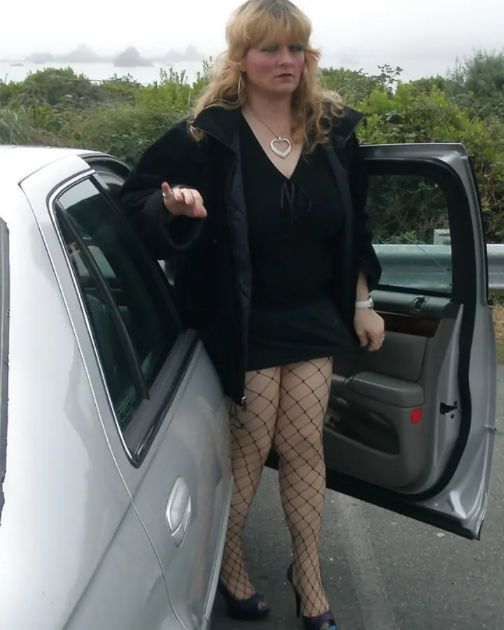 Femme blonde en robe noire, debout près d'une voiture, regardant l'appareil photo.