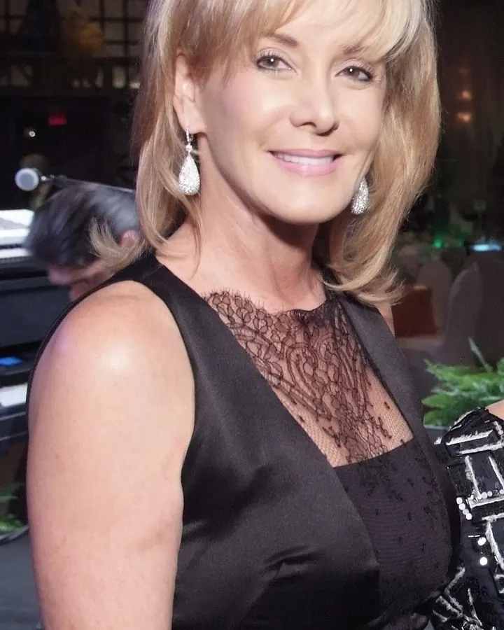 Femme d’âge mûr aux cheveux blonds courts, souriante, portant une robe blanche à motifs noirs, debout devant une cheminée en pierre.
