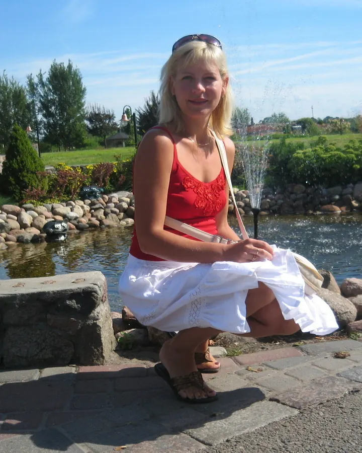 Femme blonde, souriante, portant une robe rouge et une jupe blanche, assise près d’un étang avec des jets d’eau.
