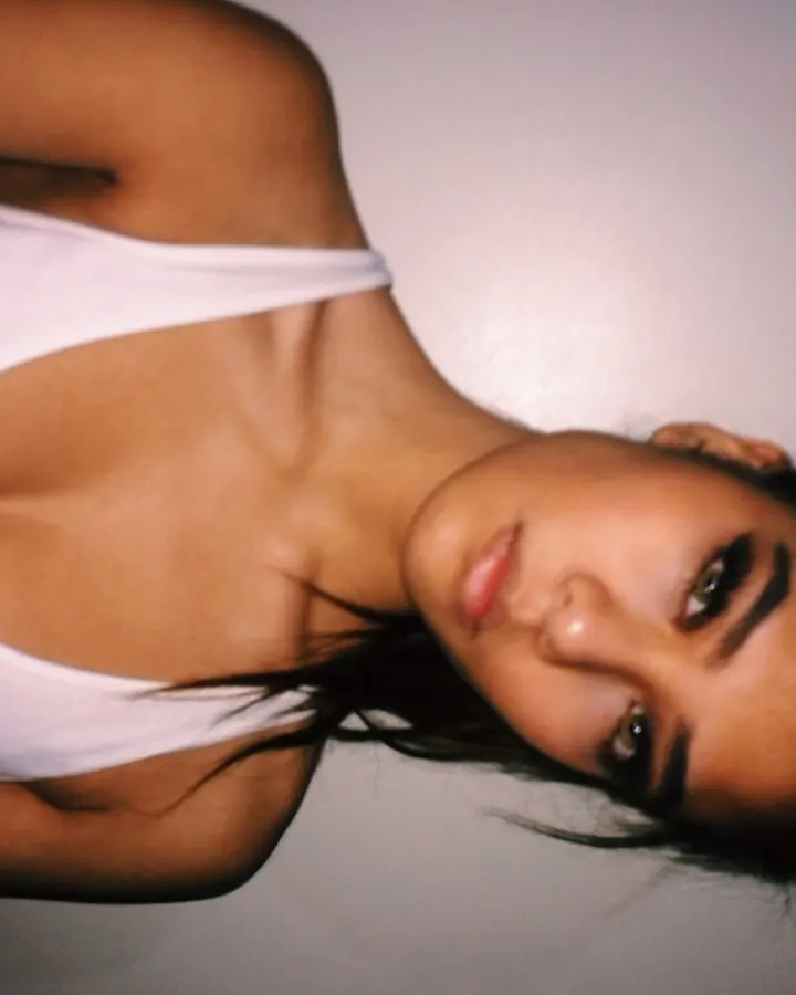Femme arabe aux cheveux bruns, en haut blanc, posant sur un fond gris, regardant l'objectif