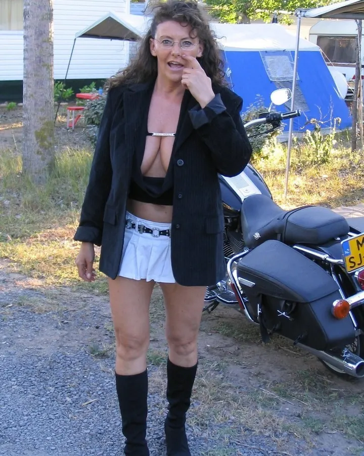 Femme mûre aux cheveux châtains, portant une veste noire ouverte sur un haut noir, debout près d'une moto