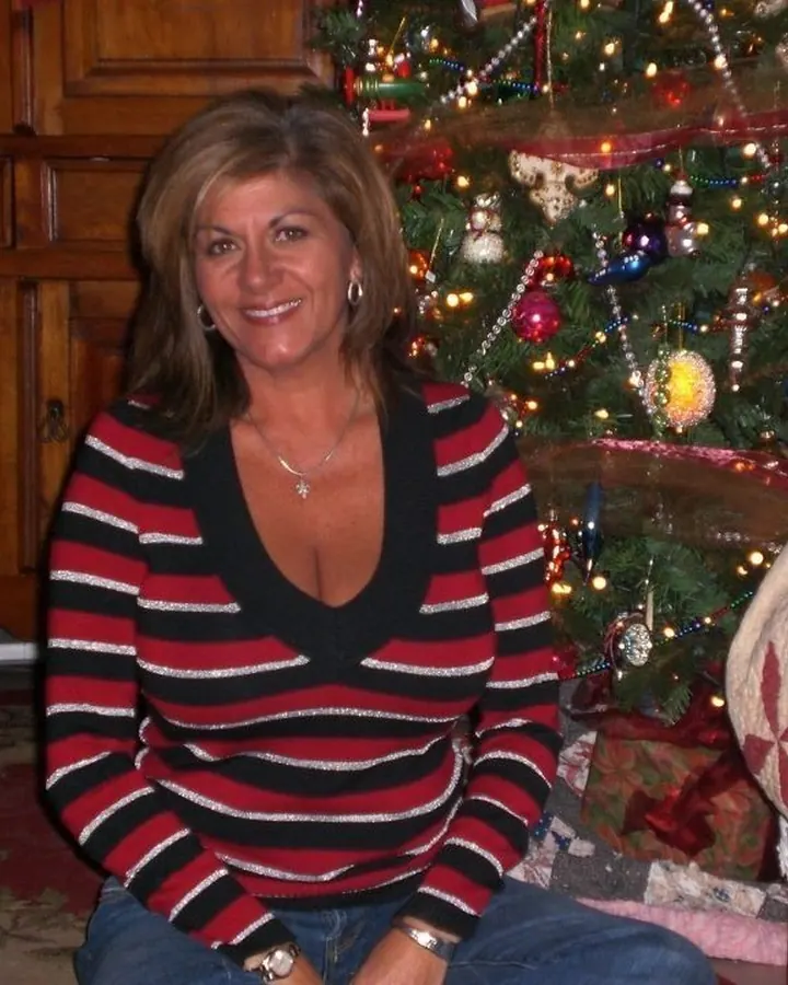Femme aux cheveux bruns portant un pull rayé rouge, noir et blanc, assise devant un sapin de Noël.