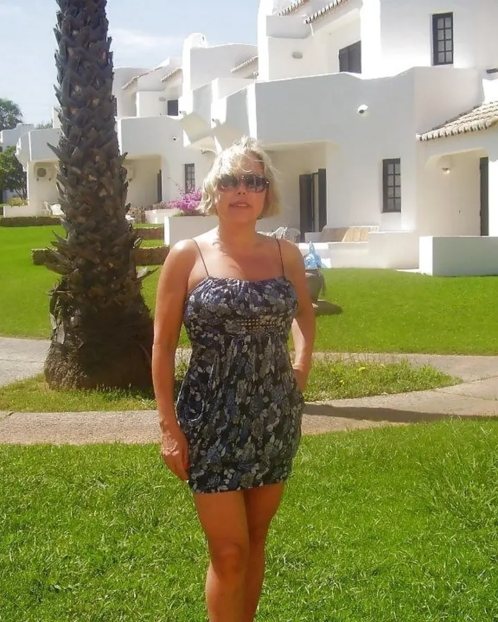 Femme aux cheveux blonds en haut noir, posant en extérieur.