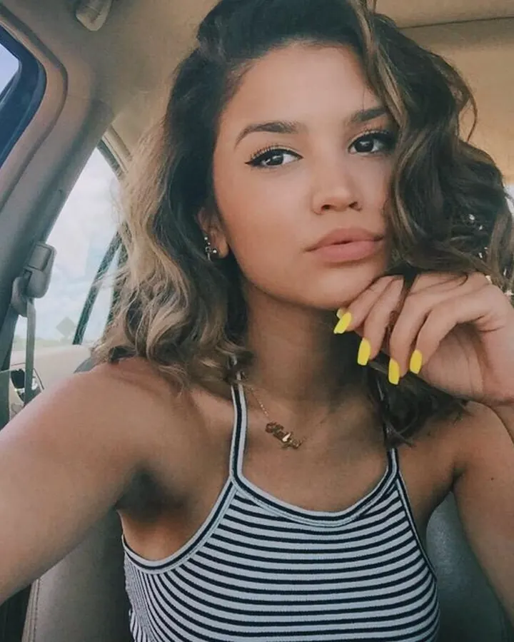 Femme aux cheveux bruns, vue de profil, assise dans une voiture, portant un haut rayé et un maquillage léger.
