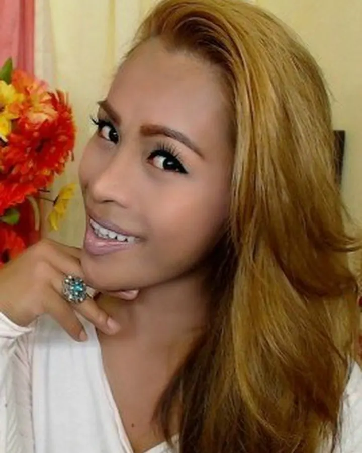 Femme aux cheveux roux, en robe rouge, debout dans un couloir, main sur la hanche, souriant.