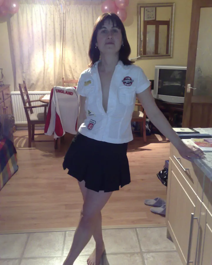 Femme aux cheveux bruns debout dans une pièce, portant une chemise blanche avec des badges et une jupe noire courte.