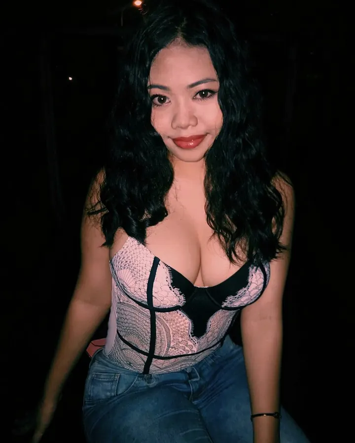 Femme asiatique aux cheveux noirs, souriante, portant un haut noir et un jean, dans un cadre sombre.
