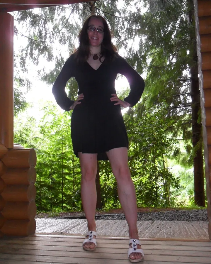 Femme en robe noire, souriante, debout sur une terrasse en bois devant une forêt verdoyante.