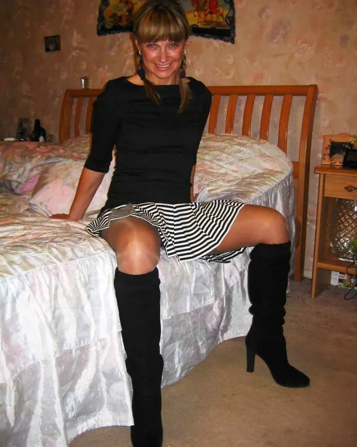 Femme en robe noire, souriante, assise sur un lit avec des bottes hautes.
