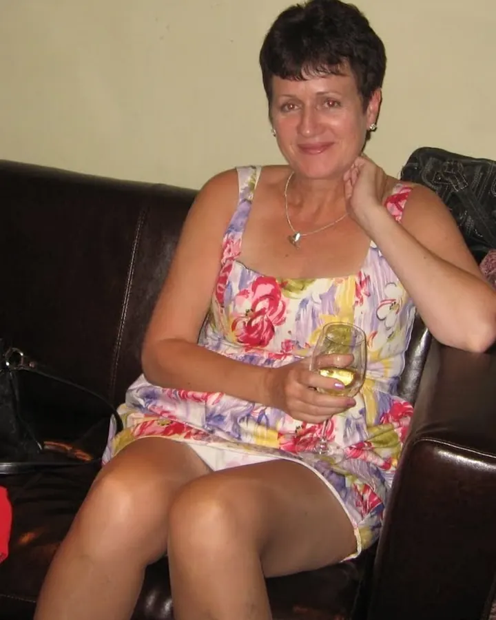 Femme aux cheveux courts, portant une robe fleurie, assise sur un canapé en cuir, tenant un verre de vin blanc, souriant légèrement.