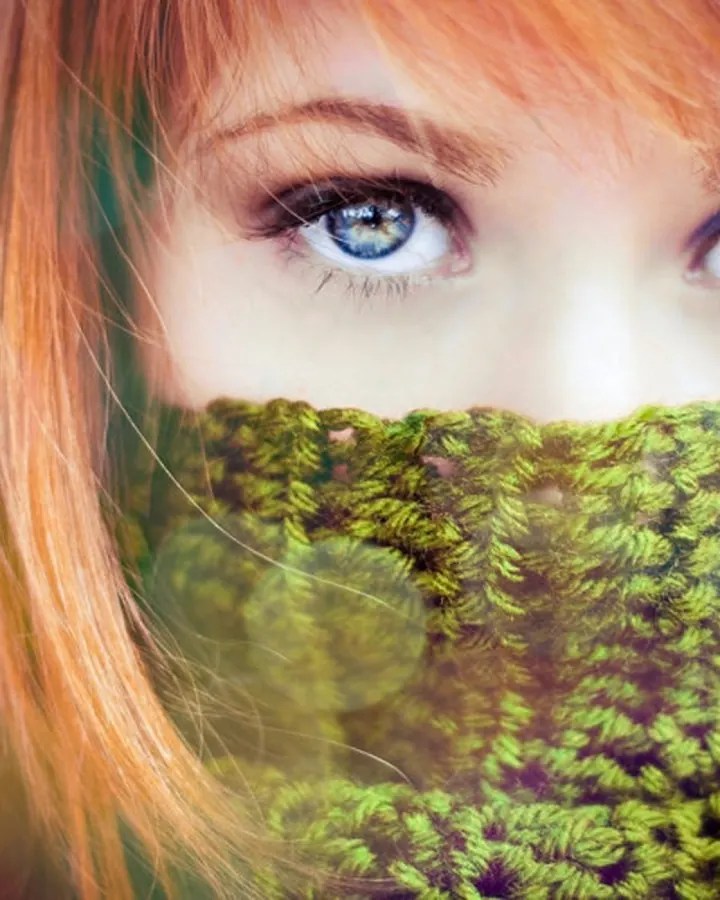 Femme aux cheveux roux et yeux bleus, regard intense, visage partiellement couvert par un foulard vert tricoté, arrière-plan flou avec des lumières colorées.