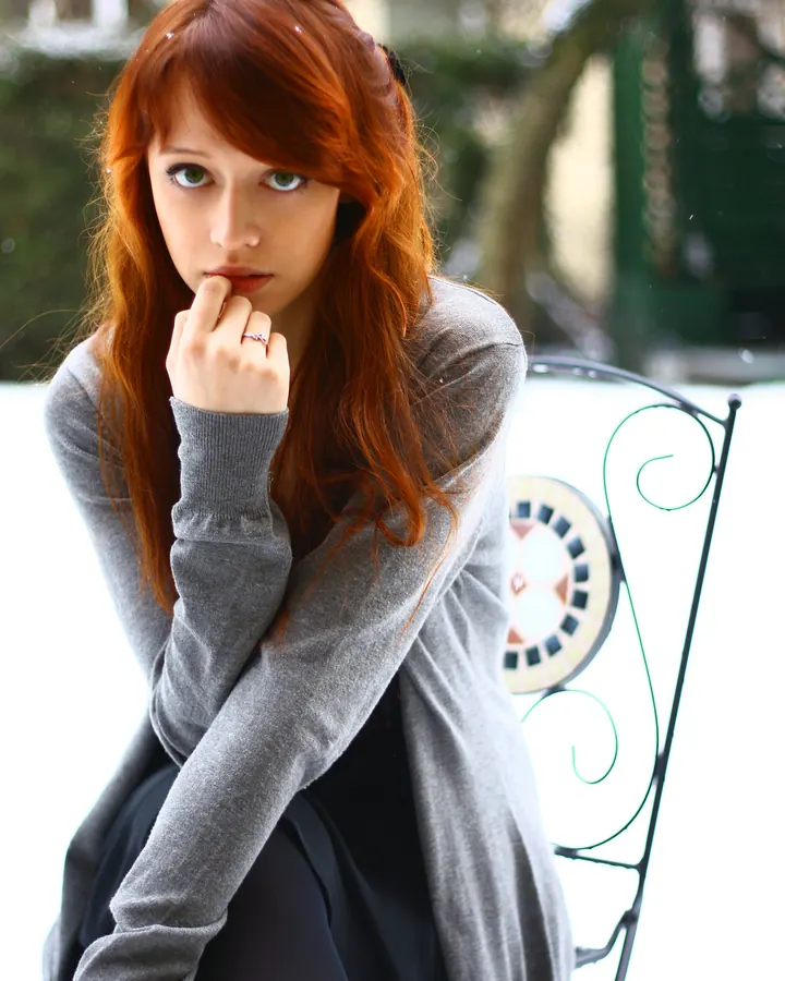 Femme aux cheveux roux portant un pull gris, assise sur une chaise en fer forgé dans la neige, main sur les lèvres.