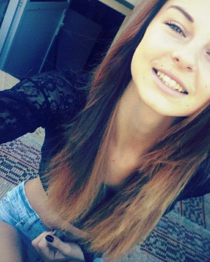 Femme aux cheveux châtains, en haut noir à dentelle, souriant à la caméra dans une pièce avec un tapis coloré.