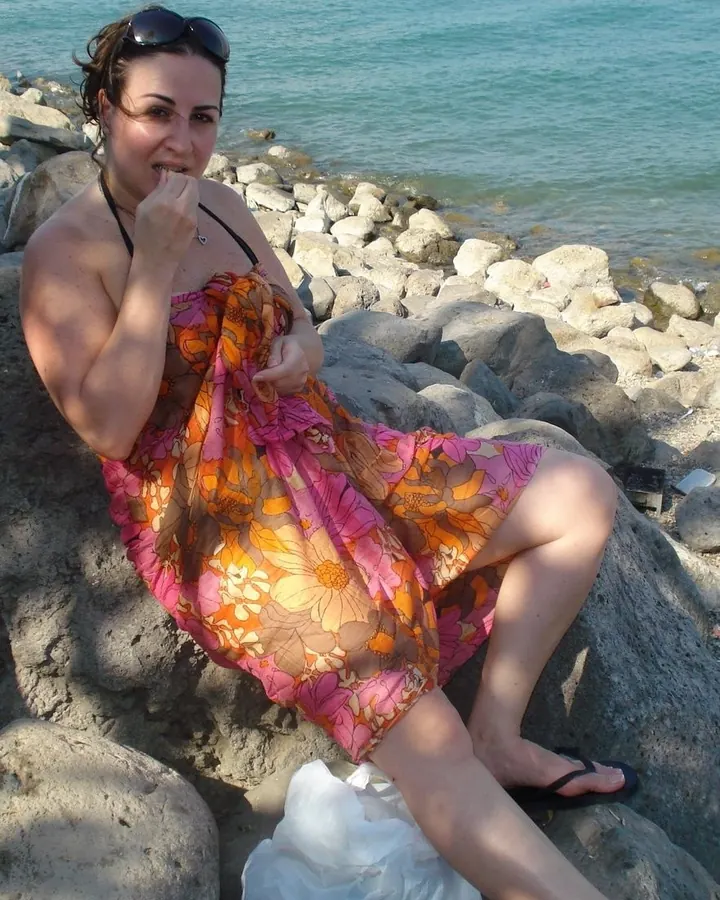 Femme assise sur des rochers près de la mer, portant une robe fleurie orange et rose, lunettes de soleil sur la tête.