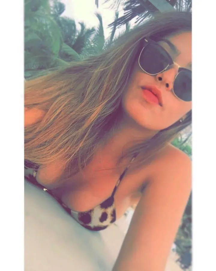Femme en maillot de bain léopard, lunettes de soleil, cheveux longs, en extérieur avec végétation.