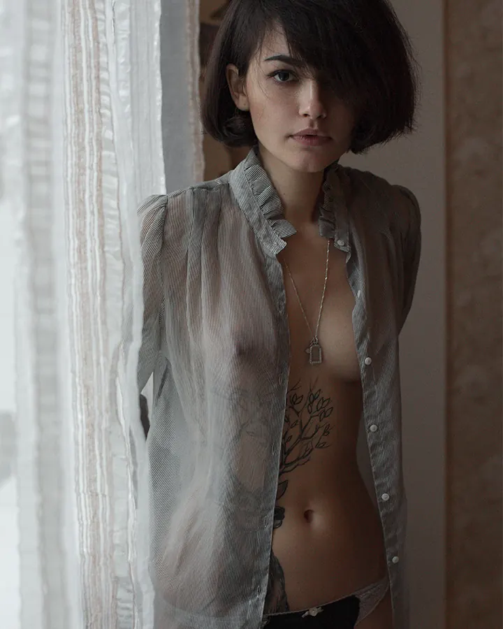 Femme aux cheveux châtains portant un haut noir, assise sur un lit, regardant l'appareil photo avec un sourire discret.