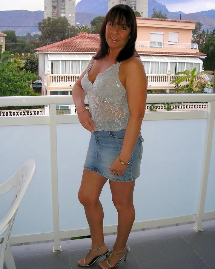 Femme aux cheveux châtains portant une chemise blanche transparente, avec un tatouage sur le ventre, debout près d'une fenêtre.