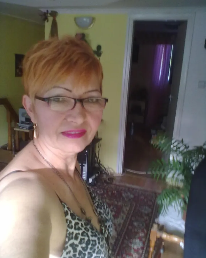 Femme aux cheveux roux portant un haut noir, assise sur un lit, regardant l'appareil photo avec un sourire discret.