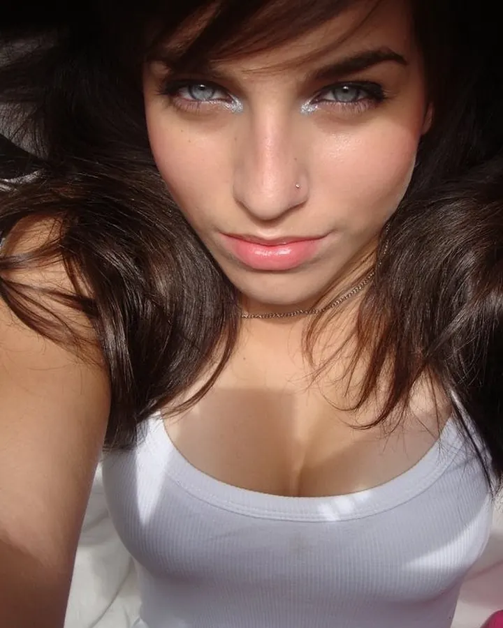 Jeune femme aux yeux bleus et cheveux châtains, en débardeur blanc, regardant l'appareil avec un sourire discret.