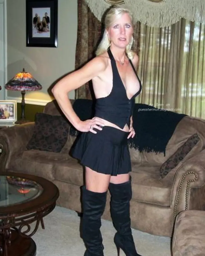 Femme blonde aux yeux bleus, debout dans un salon, en robe noire plissée et bottes hautes, mains sur les hanches.