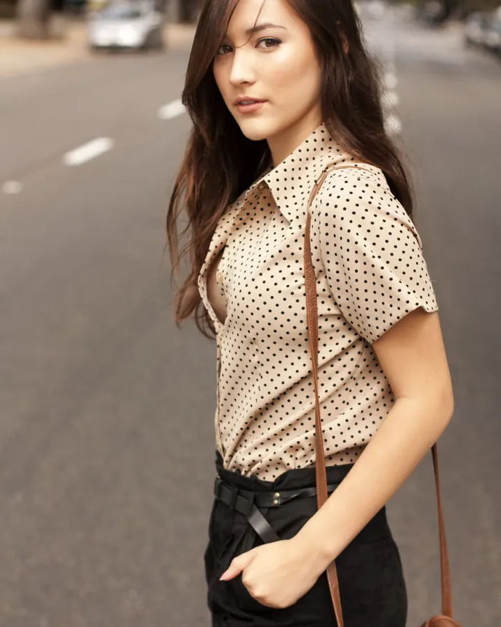 Jeune femme aux yeux marrons et cheveux châtains, en haut blanc, posant sur une rue, sac à main marron.
