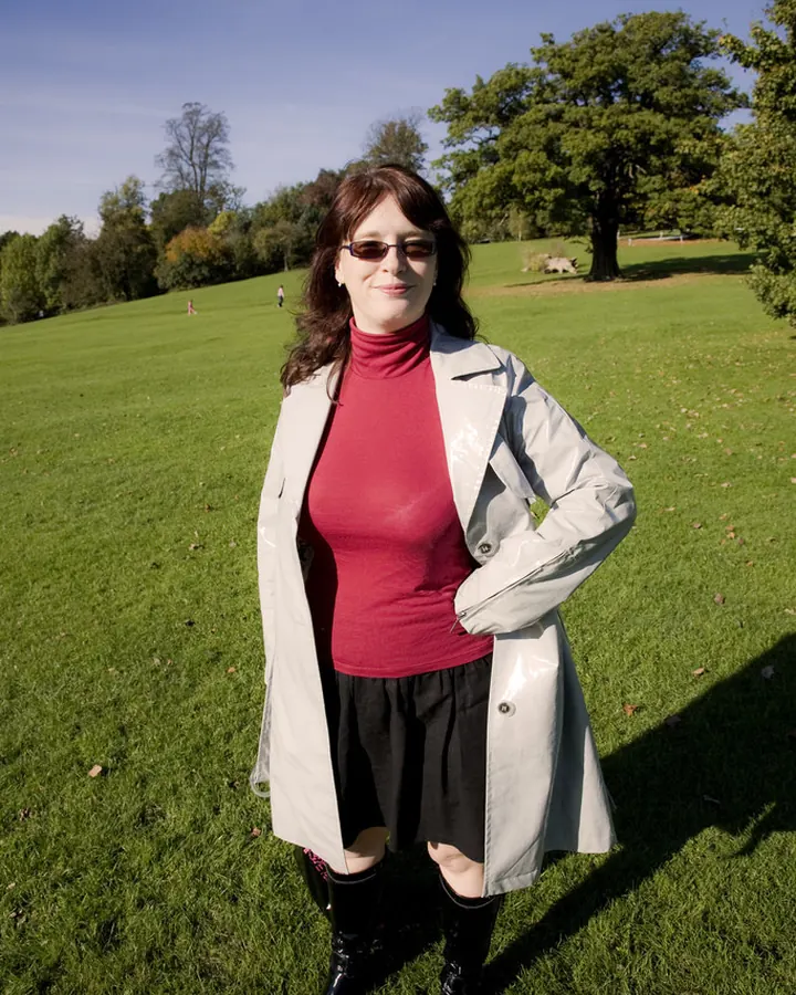 Femme en pull rouge et veste grise, debout dans un parc verdoyant sous un ciel bleu