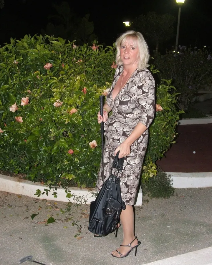 Femme blonde en robe imprimée fleurie, tenant une canne et un sac à main noir, debout près d'un buisson de fleurs roses