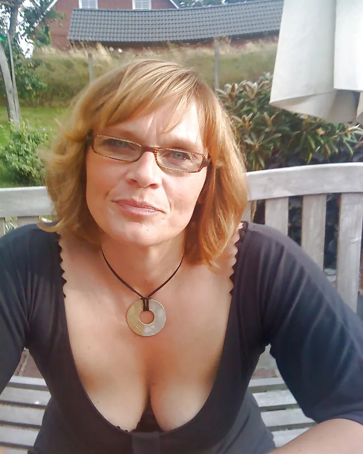 Femme blonde aux lunettes, souriant doucement, en extérieur sur une terrasse, vêtue d'un haut noir profond.
