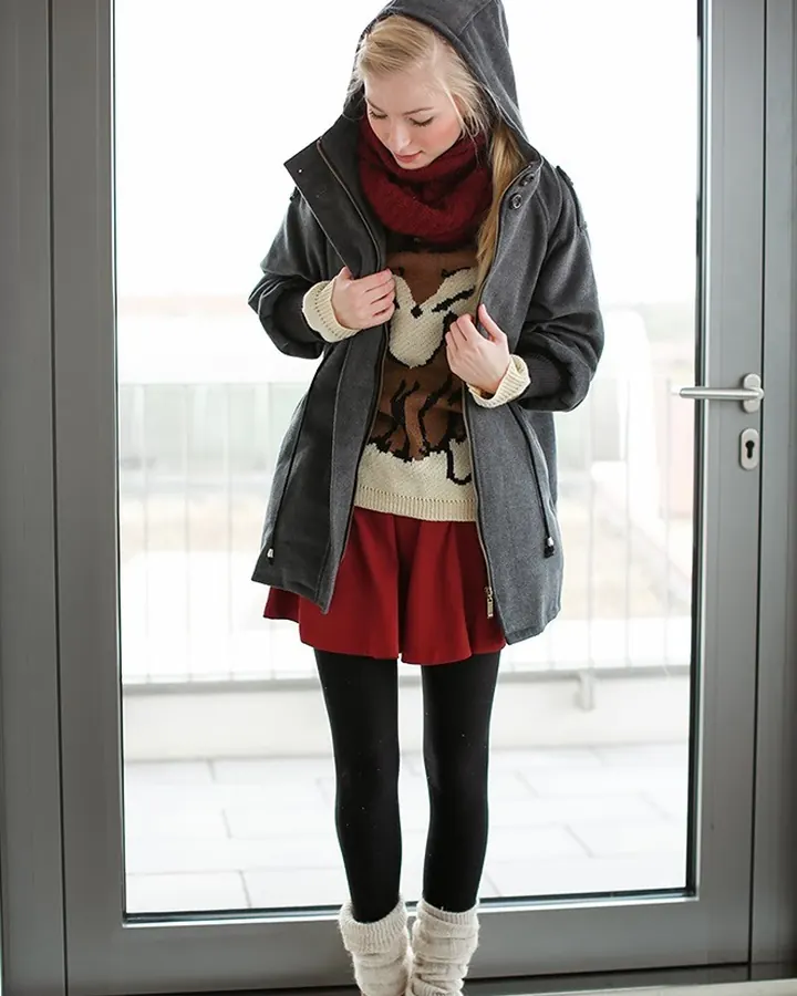 Femme aux cheveux blancs, en tenue d'hiver, debout devant une porte vitrée, tenant son pull.