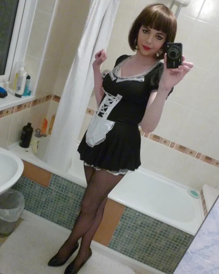 Femme aux cheveux bruns courts, portant une robe noire avec lacets blancs et un tablier blanc, des collants noirs et des chaussures noires, prenant un selfie dans une salle de bain.