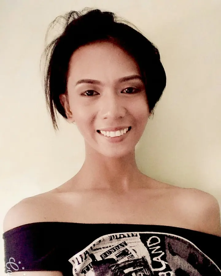 Femme aux cheveux noirs courts, souriant largement, portant un haut noir décolleté, devant un mur clair.
